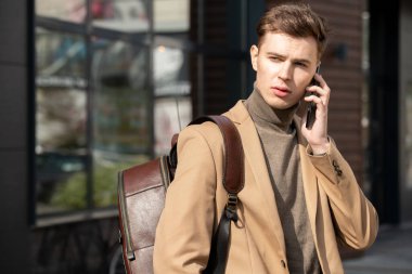 Bej ceketli ve gri kazaklı genç zarif iş adamı şehir ortamında modern mimariye karşı akıllı telefondan konuşuyor.