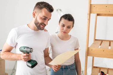 Ciddi genç sakallı adam bacak bacak üstüne atarak oturuyor ve yeni dairede mobilya montajı yaparken ders okuyor.