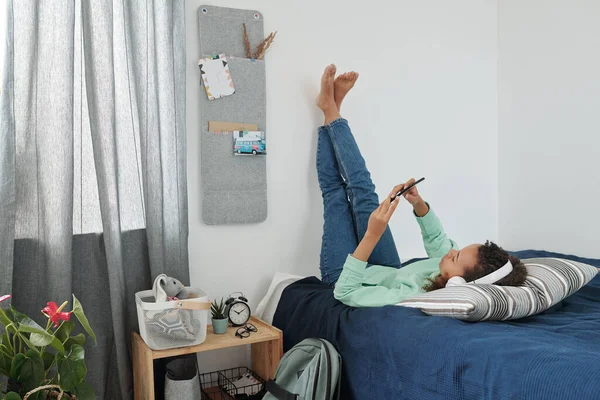 Rahatlamış Kulaklıklı Rahat Giyimli Akıllı Telefonlu Yatakta Bacaklarıyla Duvara Yaslanmış — Stok fotoğraf