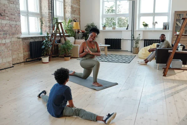 身穿灰色运动服的活泼的非洲女青年一边坐在客厅地板上的垫子上锻炼 一边向坐在绳子上的小儿子欢呼 — 图库照片