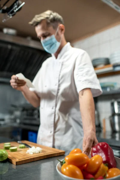 穿着制服和防护面罩的当代年轻厨师 一边拿着装有新鲜辣椒的碗 一边翻阅沙拉配料清单 — 图库照片