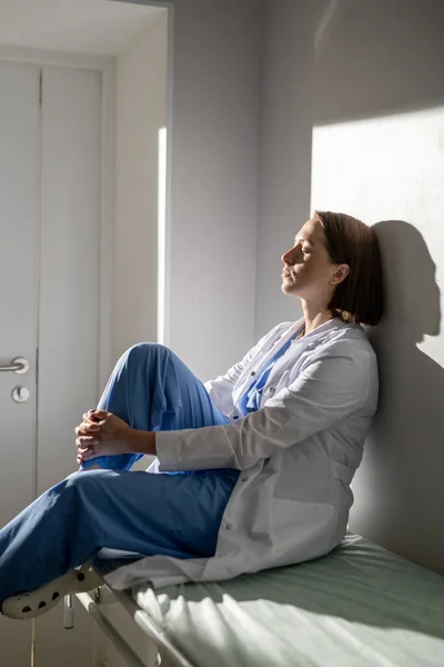 Young exhausted nurse of covid hospital in uniform sitting on medical cart and leaning against wall of chamber by window during short break