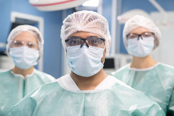 手术室里站着身穿防护服 戴面具 眼镜和工作服的年轻外科医生和他的两名助手 — 图库照片