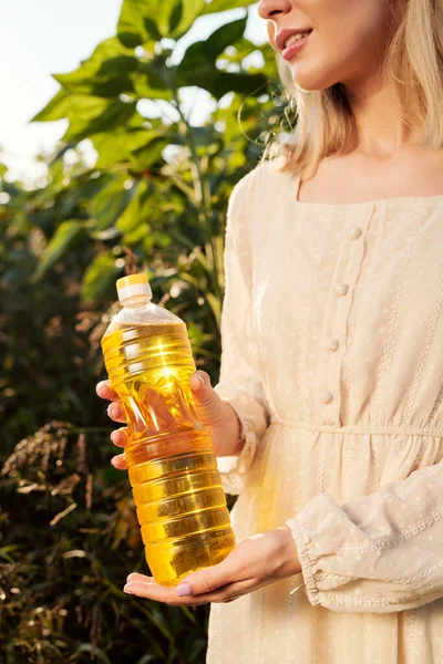 白いドレスを着た金髪の若い女性が 畑のカメラの前で緑の葉に向かって立っている間 ひまわり油のボトルを持っています — ストック写真
