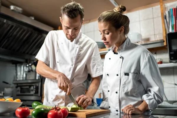 年轻的专业厨师 带着小刀 一边在餐厅厨房边吃饭 一边教他的女见习生如何煮新鲜的西葫芦 — 图库照片