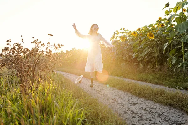 ひまわり畑の間を移動しながら楽しい時間を過ごし 晴れた日を楽しむ白いドレスの陽気な若い女性 — ストック写真