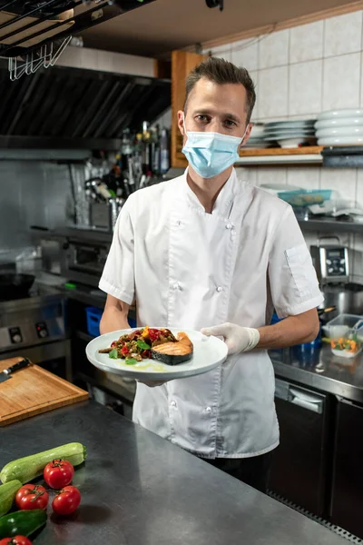 白い制服を着た若い男性シェフとキッチンテーブルの上に蒸し刻んだ野菜と揚げサーモンを保持保護マスク — ストック写真