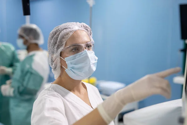 外科医生的年轻女助手 戴着手套 面罩和制服 指着医疗设备的展示 同时控制病人数据 — 图库照片