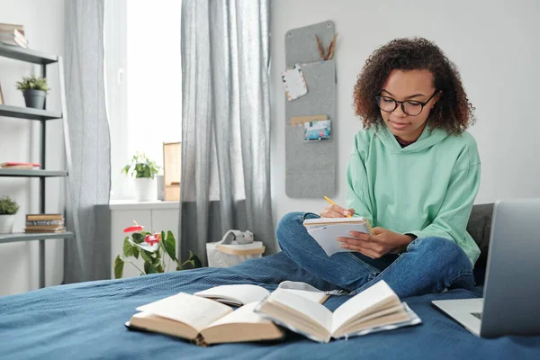 Young mixed-race diligent student in casualwear sitting on bed in front of open books and making notes in copybook in home environment