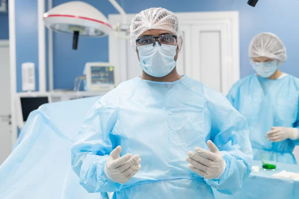 当代杂族男性外科医生 身穿防护服 戴口罩 戴手套 戴眼镜 在诊所与他的助手对视 — 图库照片