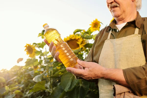 Ανώτερος Αγρότης Ποδιά Και Πουκάμισο Κοιτάζοντας Ένα Μπουκάλι Ηλιέλαιο Στα — Φωτογραφία Αρχείου