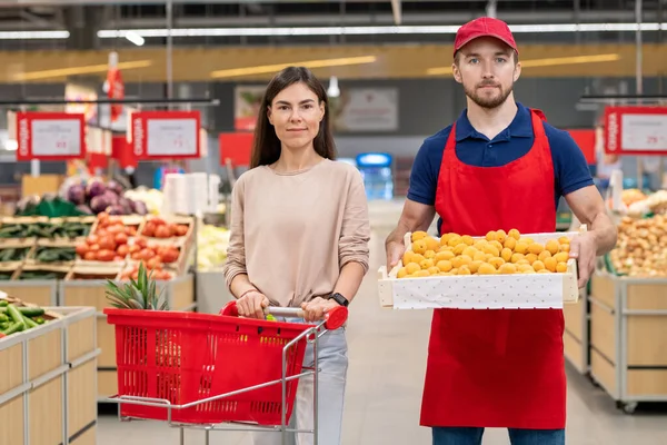 超级市场男工拿着一盒水果与年轻女性顾客站在一起的水平中长照 — 图库照片