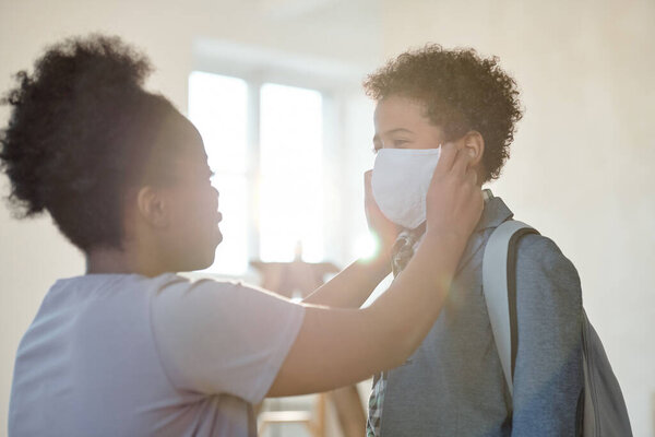 Young careful mother putting protective mask on face of her adorable little son with backpack while seeing him off to school in the morning