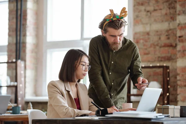 年轻而严肃的商人 带着圣诞头巾 一边指着手提电脑显示屏上的数据 一边向女同事咨询演示 — 图库照片
