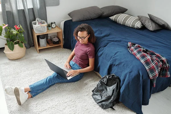 若いですリラックスした女性学生ですカジュアルウェアで床に座ってベッドでラップトップで足の上に家の環境でネットサーフィンしながら — ストック写真
