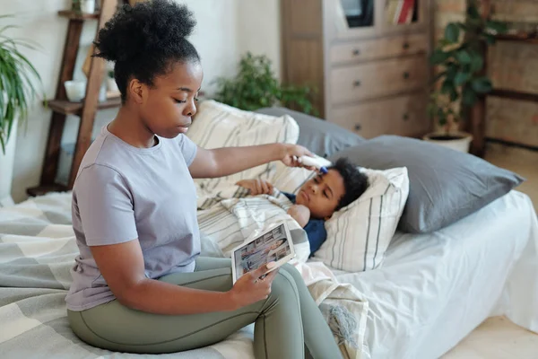 年轻的非洲妇女在床上用毯子测量她生病的小儿子的体温 并观看在线医疗救助视频 — 图库照片