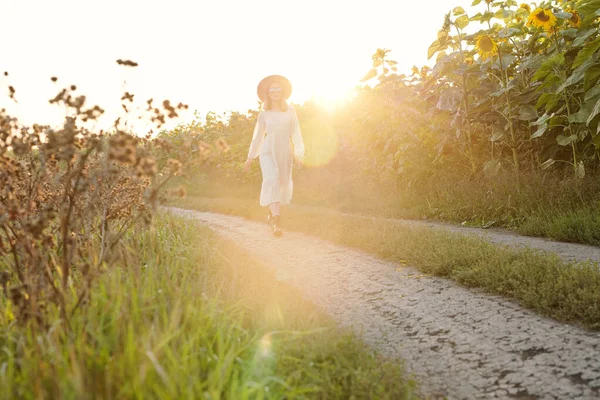 戴着帽子 戴着太阳镜 穿着衣服的漂亮姑娘走在向日葵地之间的人行道上 在阳光灿烂的早晨享受着散步 — 图库照片