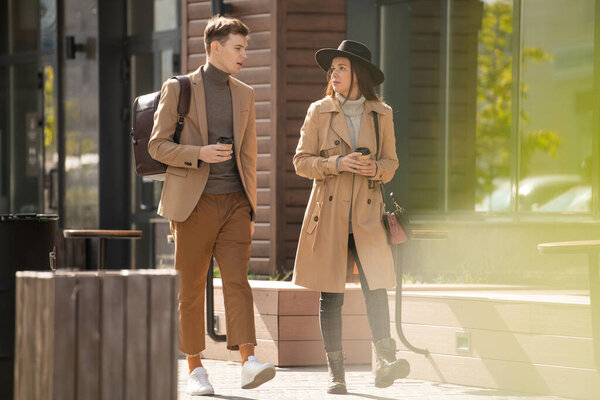 Young serious stylish woman with drink talking to her boyfriend in casualwear walking near by in urban environment during chill