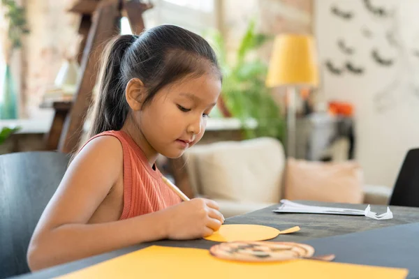 聚精会神的亚洲女孩坐在桌旁 在艺术工作室为万圣节做南瓜纸制工艺 — 图库照片