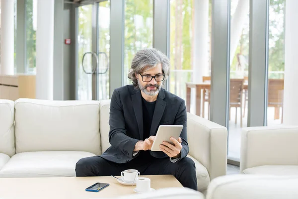 Bearded mature elegant banker in suit and eyeglasses scrolling through online financial data in tablet by cup of coffee in restaurant