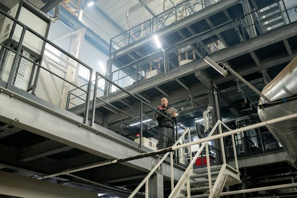 階段の前に立っている間 現代的な大規模なポリマー加工工場内で働くデジタルタブレットと大胆な成熟したフォアマン — ストック写真