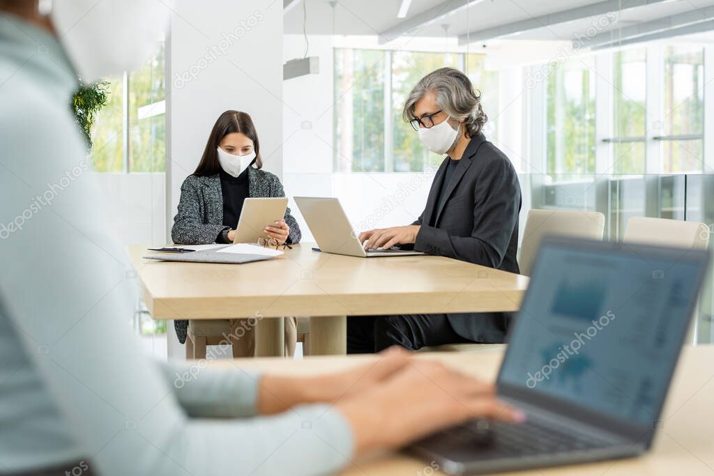 Two busy colleagues in smart casualwear and protective masks sitting by table and using mobile gagdets while networking in large office