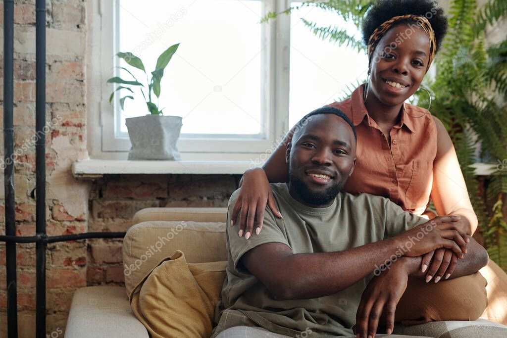 Joyful young African couple in casualwear looking at you with smiles while sitting in front of camera against window with domestic plants