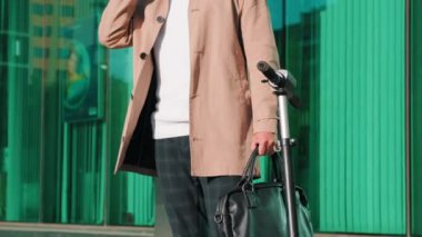 Siyah güneş gözlüklü, şık iş adamının deri çantalı ve telefonda modern ofis binasının yanında e-scooterlı bir şekilde konuşurken çekilen ayak parmaklarından yukarı doğru eğik pozlarına kadar.