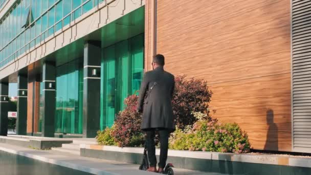 バックビューサングラスとコートを着た大人の男の完全な映像晴れた日に市内中心部の近代的なオフィスビルに沿って電動スクーターで歩く — ストック動画