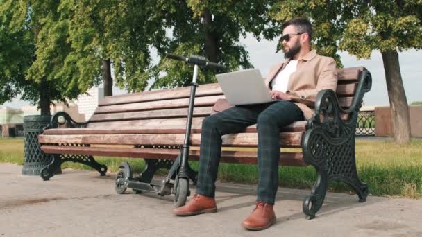 全景拍摄的现代商人坐在公园的长椅上 带着电动车在笔记本电脑上工作 并在日光下环视四周 — 图库视频影像