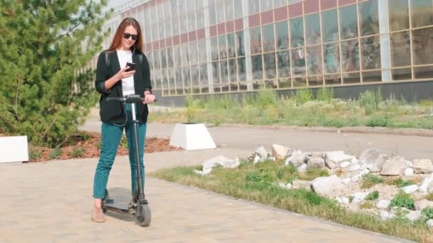 在公园里站在她的现代电子摩托车旁边 手持智能手机的年轻女子的全照 — 图库视频影像