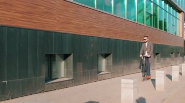Şık giyinmiş, güneş gözlüklü, elektrikli scooterlı camdan ofis binası boyunca ayakta duran bir adam.