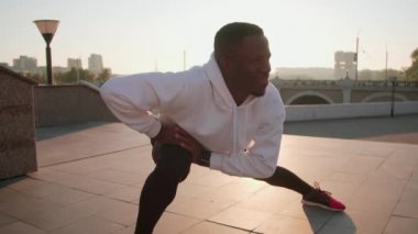 Spor giyim kuşamında, neşeli zenci bir adam sabah antrenmanında gülümseyen ve dışarıda eğlenen bir bayan arkadaşla yan hamleler yapıyor.