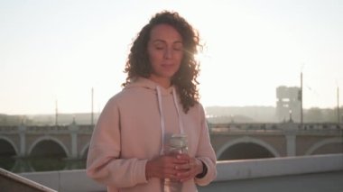 Açık hava antrenmanında elinde bir şişe su olan güzel kıvırcık saçlı bir kadının arka planda güneşli bir sabahta şehir köprüsünde kameraya gülümserkenki görüntüsü.