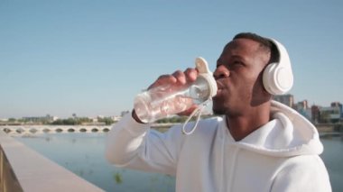 Gündüz vakti açık mavi gökyüzüne bakarak koşu yaptıktan sonra spor şişesinden su yudumlayan vahşi Afro-Amerikan adam.