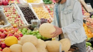 Kot ceketli sakallı genç adamın el bilgisayarı markette kavun seçiyor.