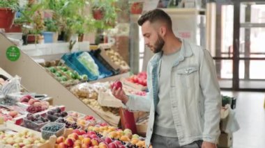 Orta boy kot ceketli sakallı bir genç adam markette meyve standına doğru yürüyor ve nektarin seçiyor.