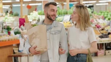 Alışveriş torbalarıyla dolu neşeli genç bir çiftin portresi markette duruyor ve kameraya gülümsüyor.