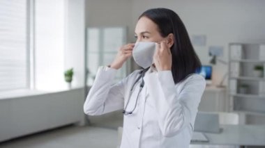 Doktorun ofisinde dikilip koruyucu maske takıp kameraya bakarken çekilmiş bir portre.