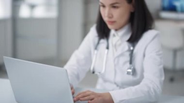 Güzel Asyalı doktorun portresi ofiste oturmuş dizüstü bilgisayarla veri üzerinde çalışırken çekildi.