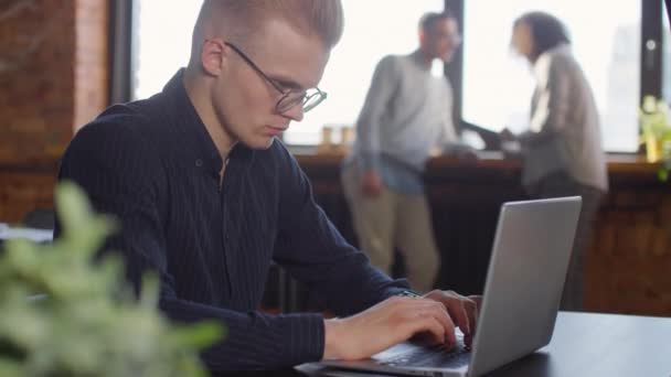 彼の同僚とノートパソコンで作業している若い白人男性の傾きアップ現代のロフトオフィスで背景にパノラマの窓で通信 — ストック動画