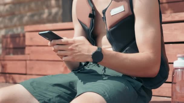 夏に屋外のベンチに座っている間 手のテキストメッセージでスマートフォンとウォータースポーツライフジャケットの認識できない男性サーファーのミッドセクション映像 — ストック動画