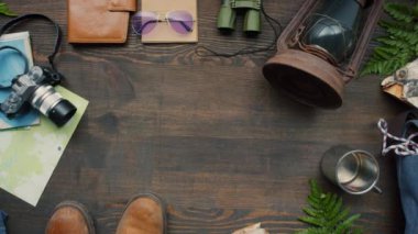 En üstteki düz görünümde yürüyüş botları, fotoğraf makinesi, halat, harita, sırt çantası, fener, defterler, seyahat kupası ve eğreltiotu ile süslenmiş ahşap masanın üzerinde duran dürbün görüntüleri var.