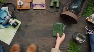 En üstte, tanınmayan bir kadının, üzerinde seyahat aksesuarları bulunan pasaportları tahta masaya koyarkenki görüntüsü var.