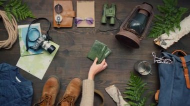 Üst manzara, tanınmayan bir kadının seyahat için bavulunu hazırlarken tahta masaya pasaport koyuşunu gösteriyor. Seyahat aksesuarları masada yatıyor.