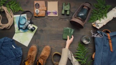 Haritalar, ip, seyahat giysileri, sırt çantası, pusula, fener, dürbün ve eğreltiotu ile süslenmiş ahşap masanın üzerinde duran fotoğraf makinesi. Masaya pasaport koyan tanınmayan bir kadın.