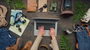 Tanınmayan bir kadının dizüstü bilgisayarda yazıp sonra da bilgisayarı kapatıp götürdüğü en iyi görüntü. Ahşap masada duran fotoğraf makinesi, harita, fener ve ip gibi çeşitli seyahat aksesuarları.