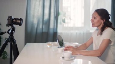 Orta yaşlı güzel bir kadının, sıcak oturma odasında otururken kameraya bakıp kendini filme çekerek gençleştirici kahve fırçası tarifi verirken orta ölçekli görüntüsü.