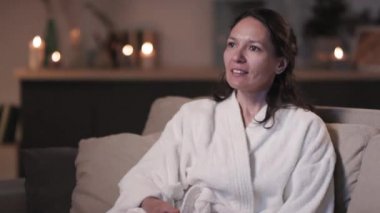 Gülümseyen, beyaz bornozlu, rahatlamış bir kadının televizyon karşısında güzellik prosedürleri ve vücut bakımından sonra televizyon kanallarını değiştirmesi.