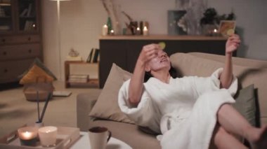 Medium shot of beautiful smiling woman with facial mask relaxing after hard day applying cucumber sticks on eyes lying on comfy sofa